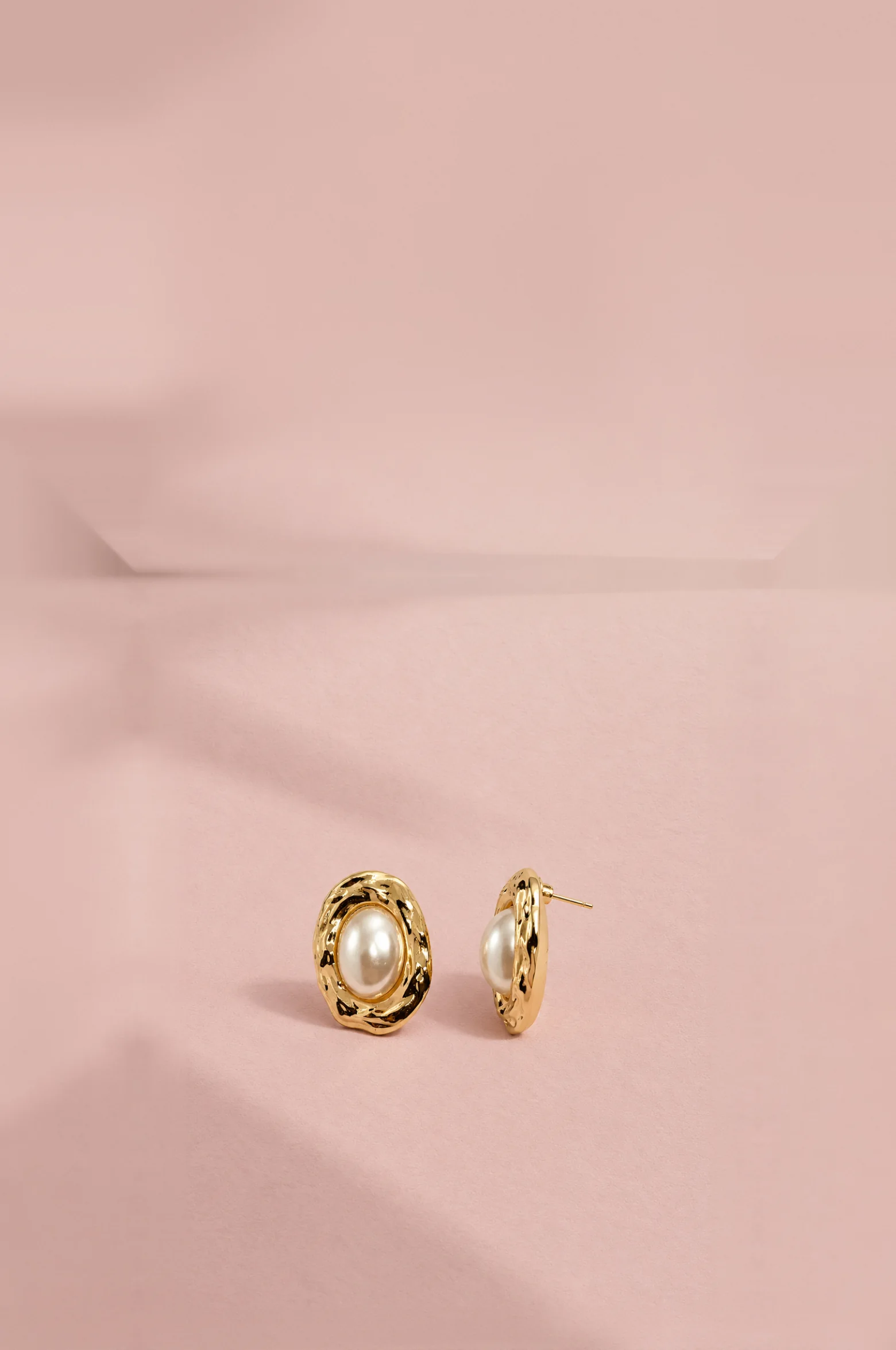 Gold-framed pearl stud earrings resting on a blush pink surface